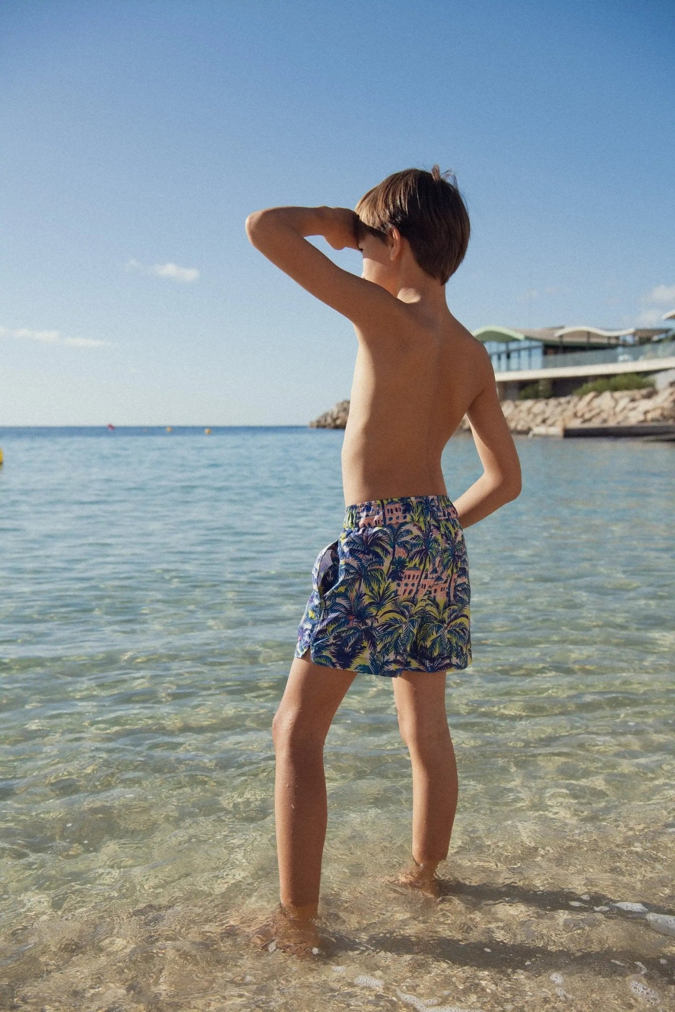 Boy in blue and gold jungle print swim shorts standing in shallow beach water