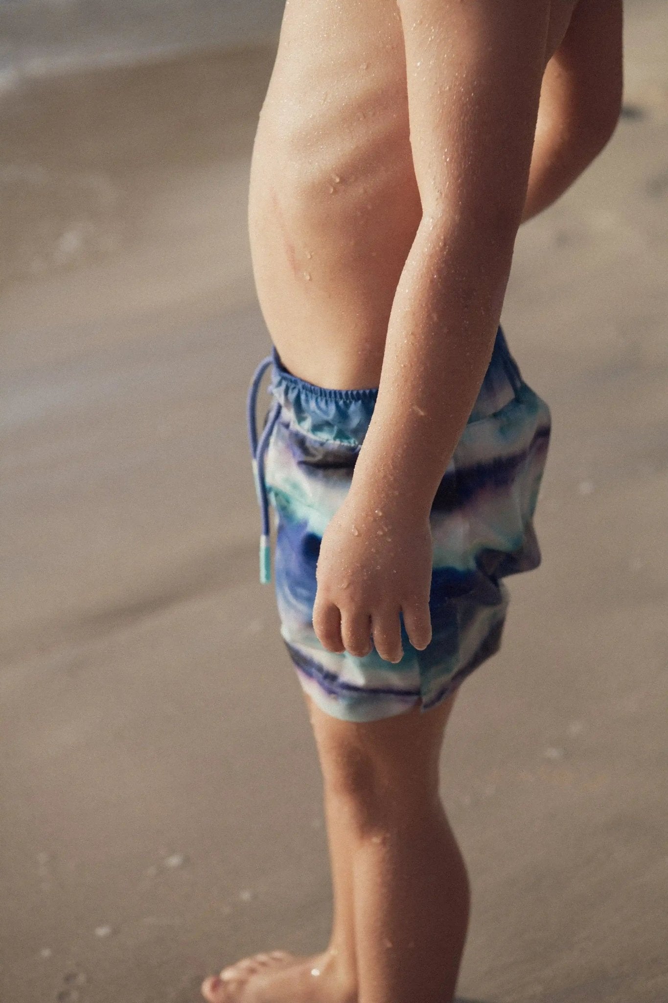 Boy wearing printed swim shorts at the beach, water droplets on skin, summer swimwear