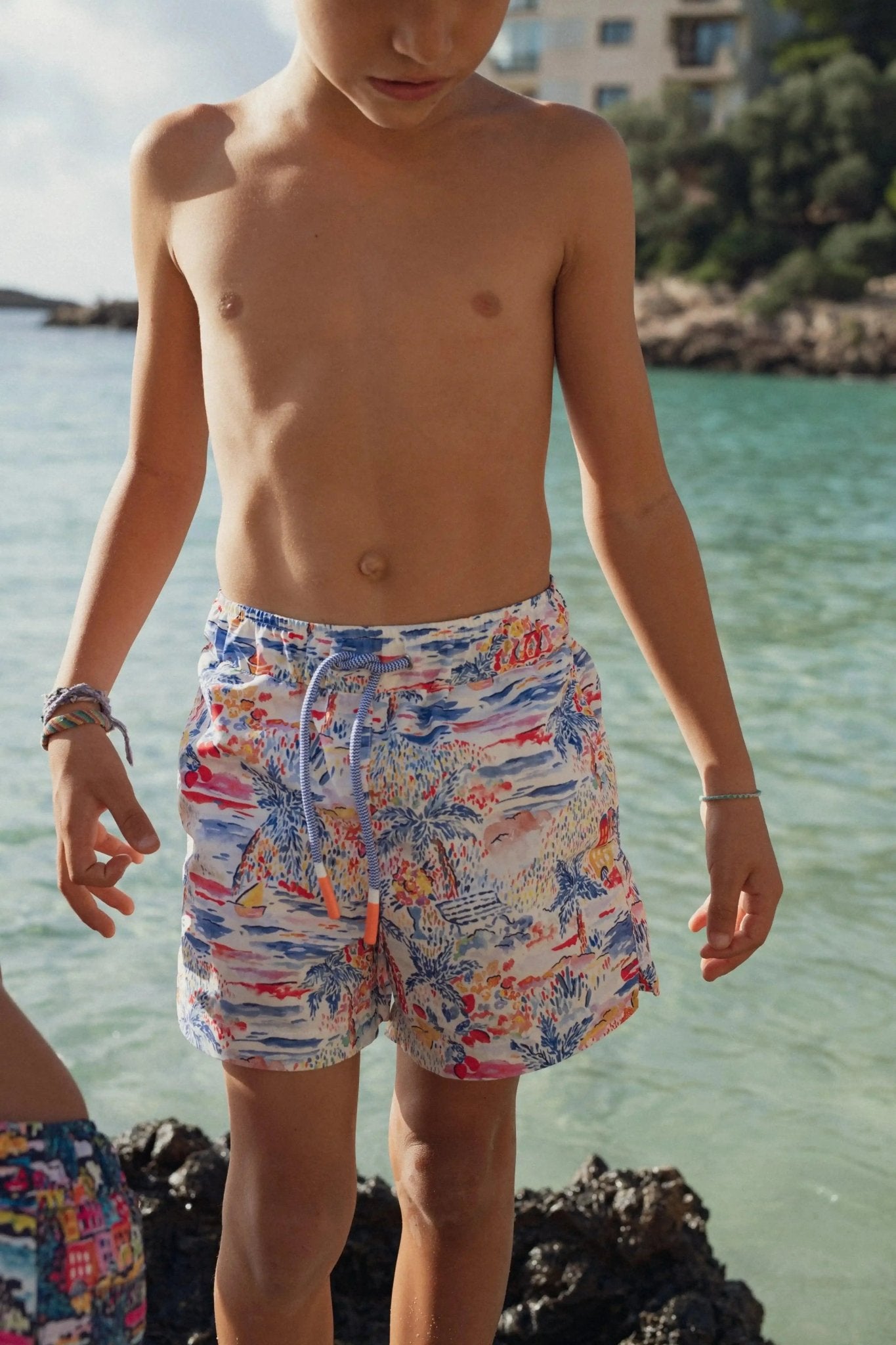 Boy wearing colorful printed swim shorts by the sea, tropical beachwear style
