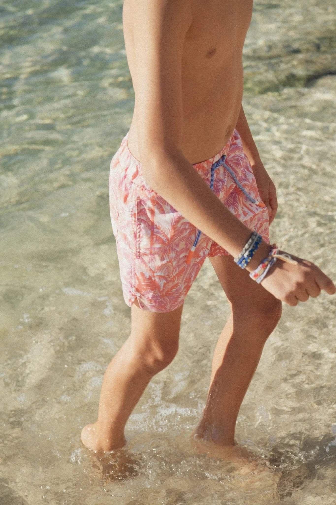 Boy wearing orange jungle print swim shorts walking in clear beach water