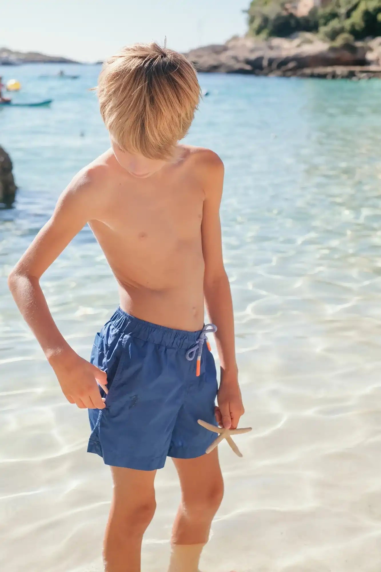 Boy wearing indigo swim shorts by Lison Paris on a sunny beach, holding a starfish.