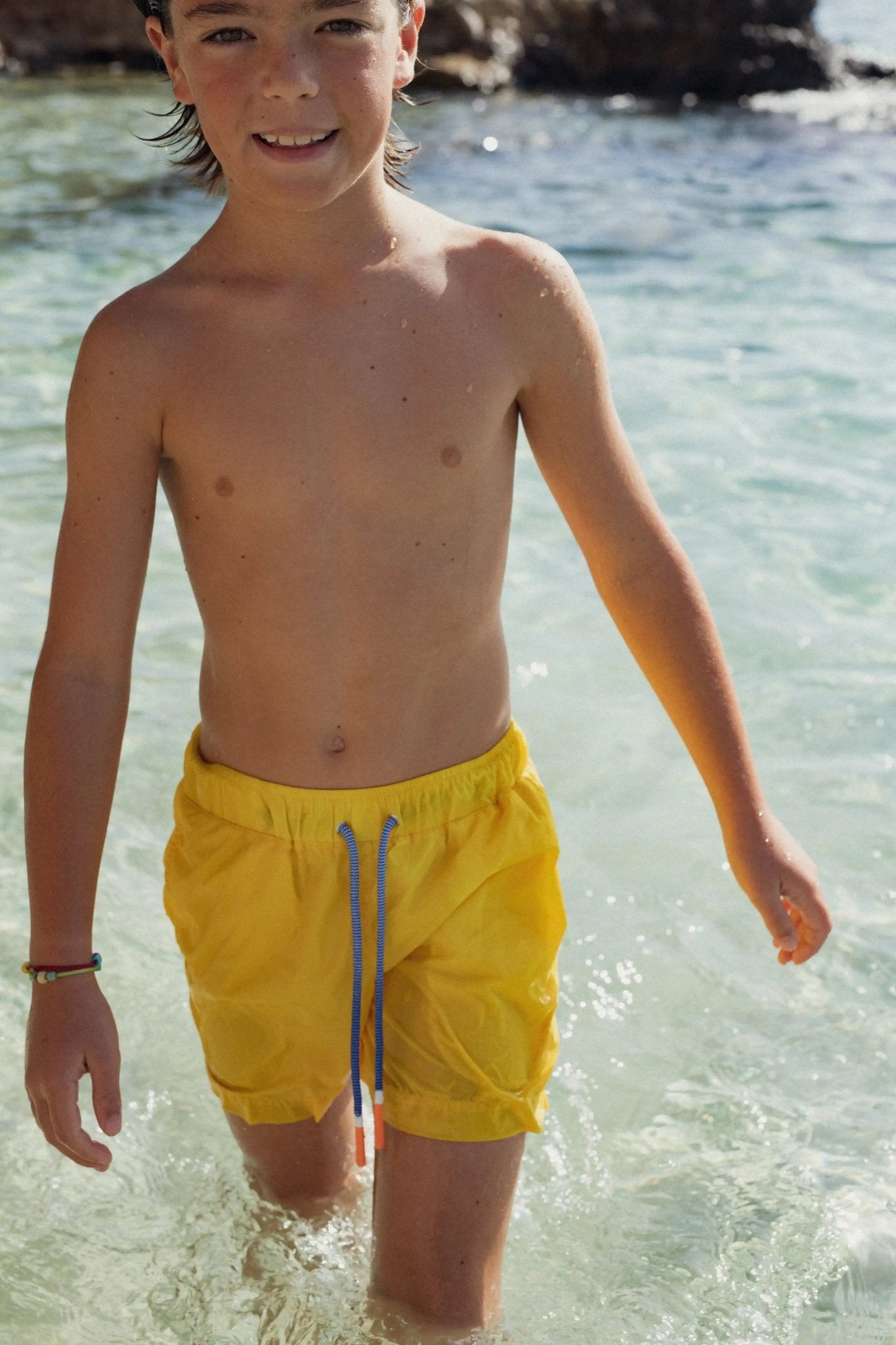 Smiling boy wearing yellow swim shorts standing in shallow clear beach water