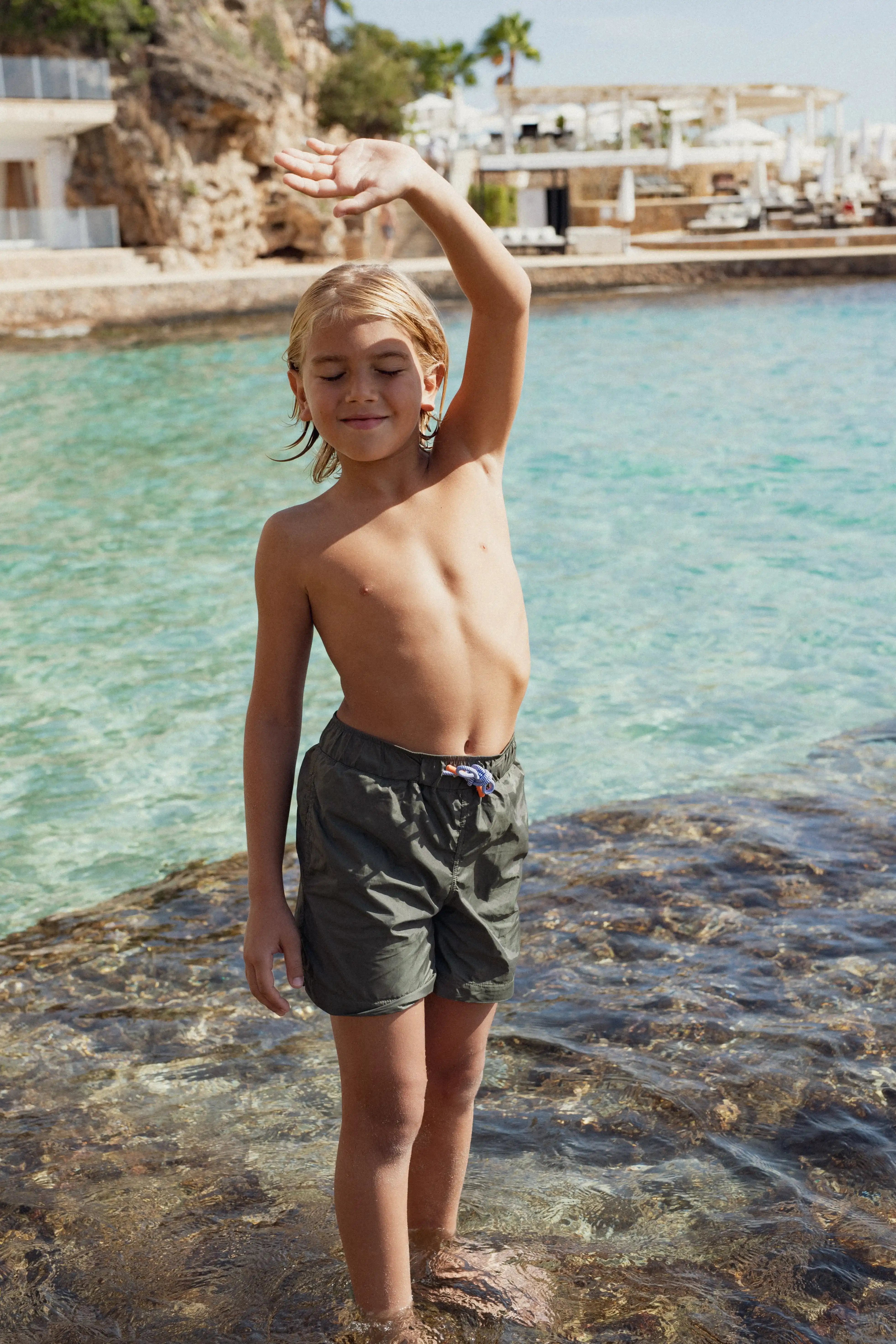 Boy wearing khaki swim shorts standing in clear water, Lison Paris beachwear summer scene
