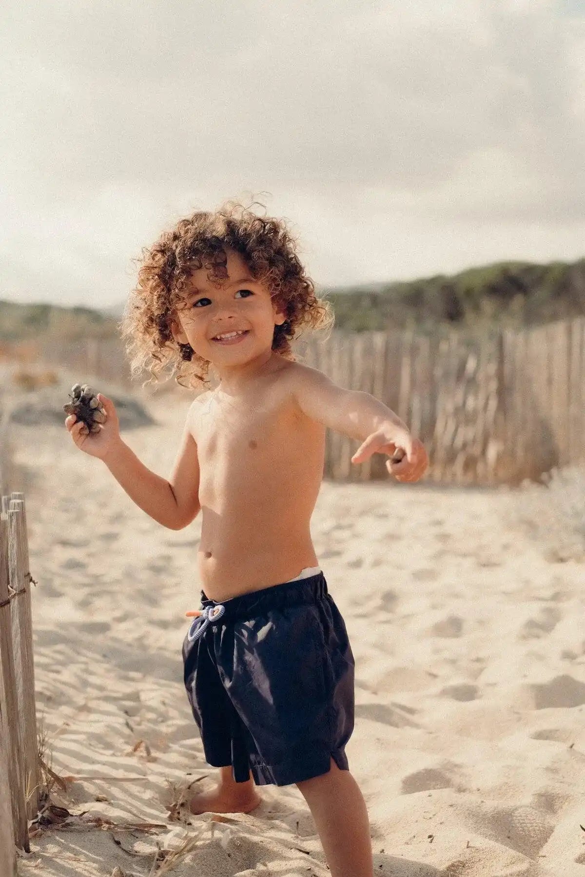 Smiling boy in navy blue swim shorts on a sunny beach, Lison Paris kids beachwear