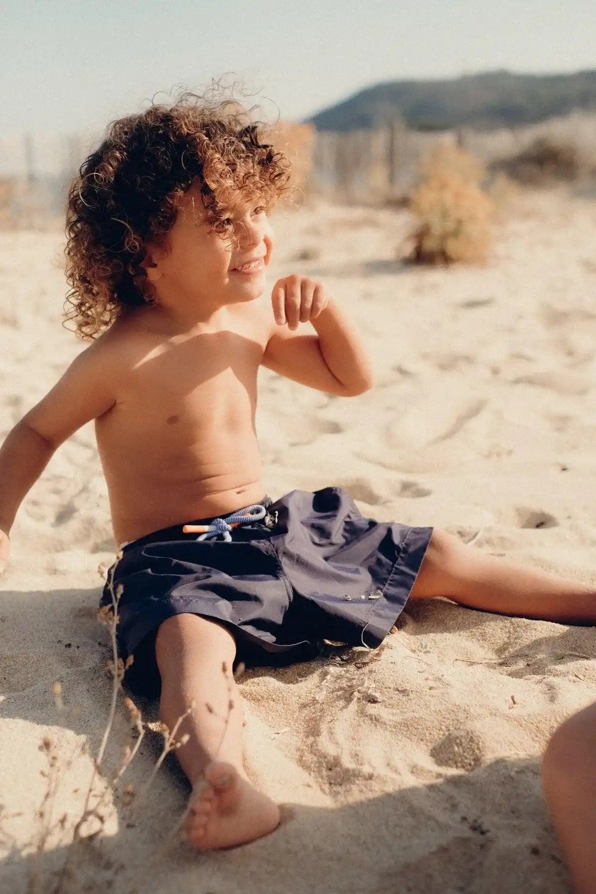 Smiling boy in navy swim shorts sits on sandy beach, Lison Paris kids beachwear