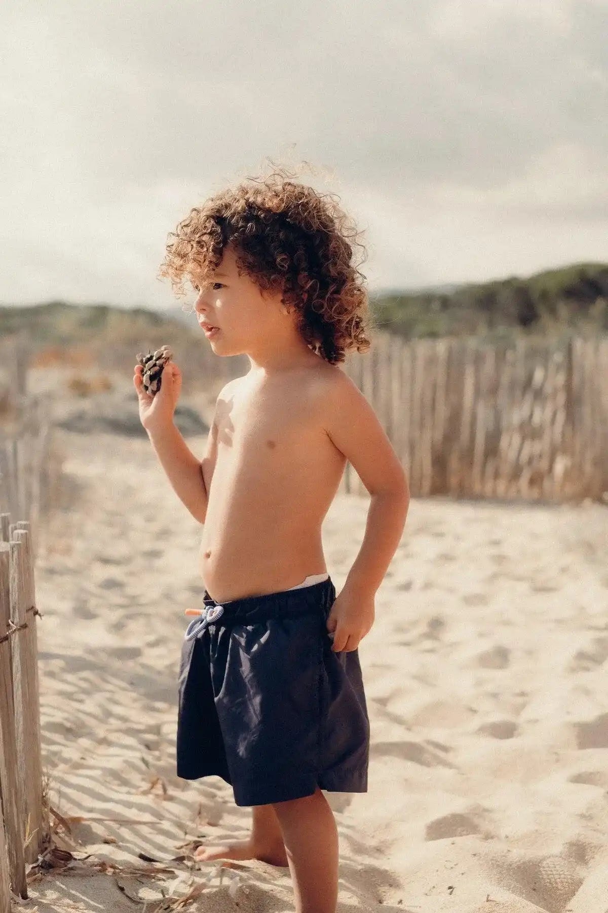 Boy in navy swim shorts on sandy beach, Lison Paris UV swimwear collection