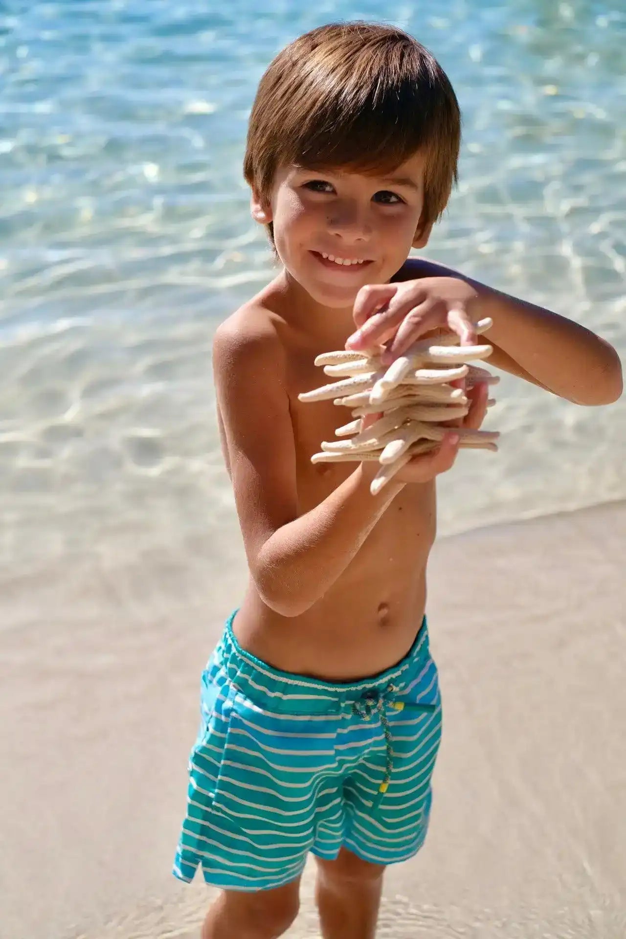 Boy in blue striped swim shorts holding starfish on a sunny beach near clear water