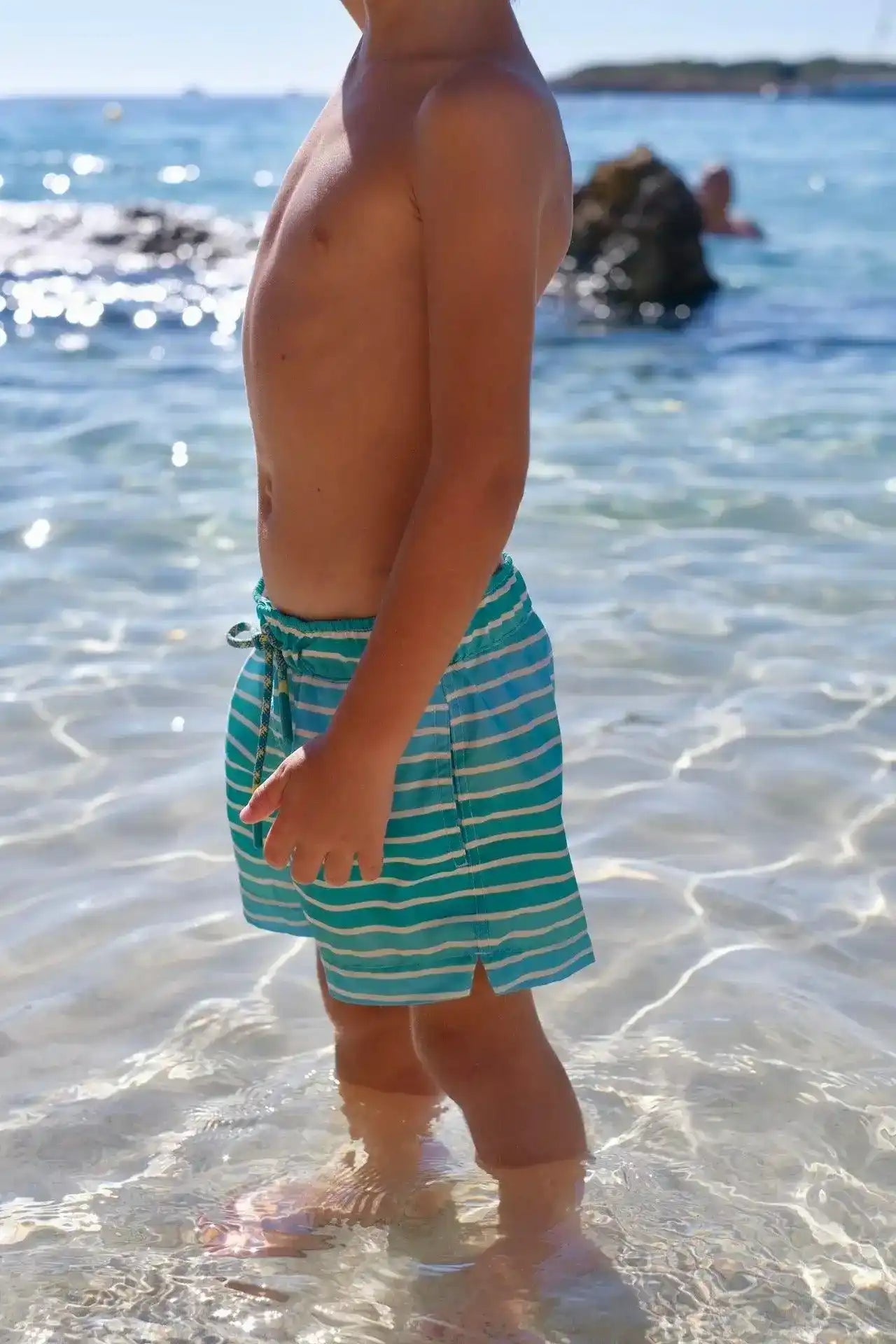 Boy wearing blue striped swim shorts standing in shallow beach water