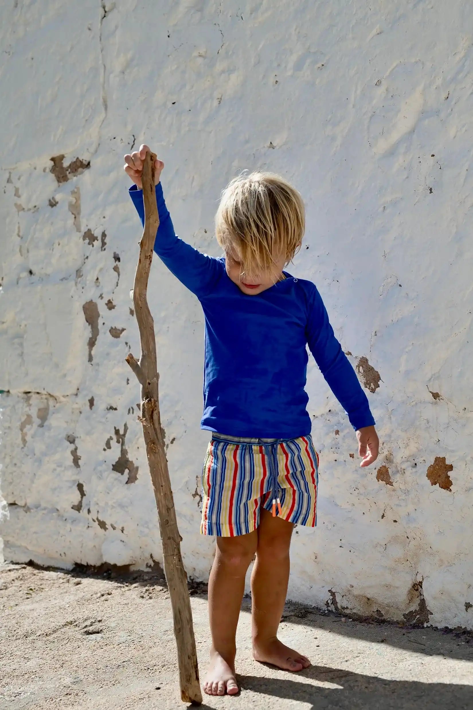 Boy wearing Lison Paris multicolored striped swim shorts and blue UV shirt outdoors
