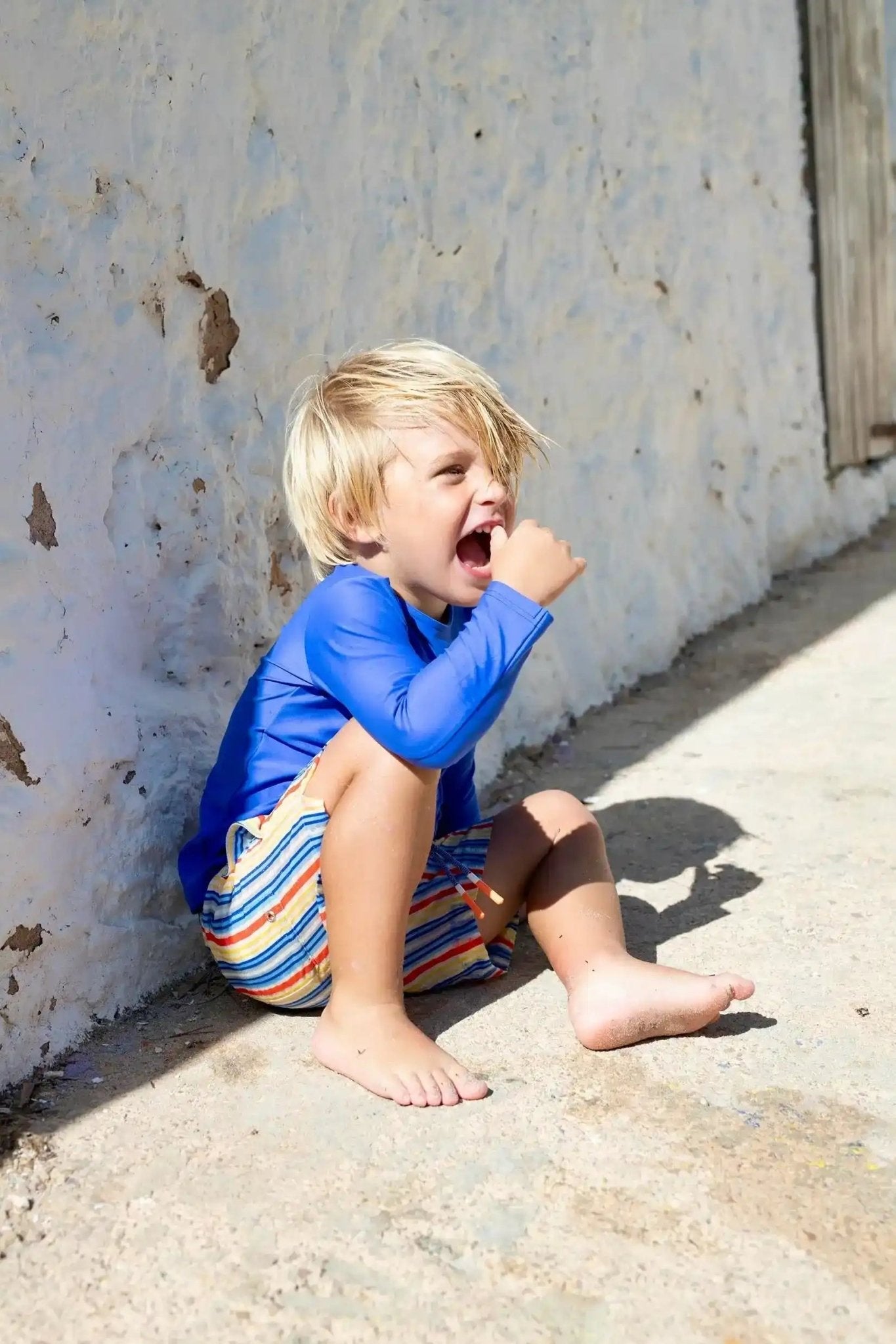 Boy in colorful striped swim shorts and blue rash guard sitting by a sunlit wall