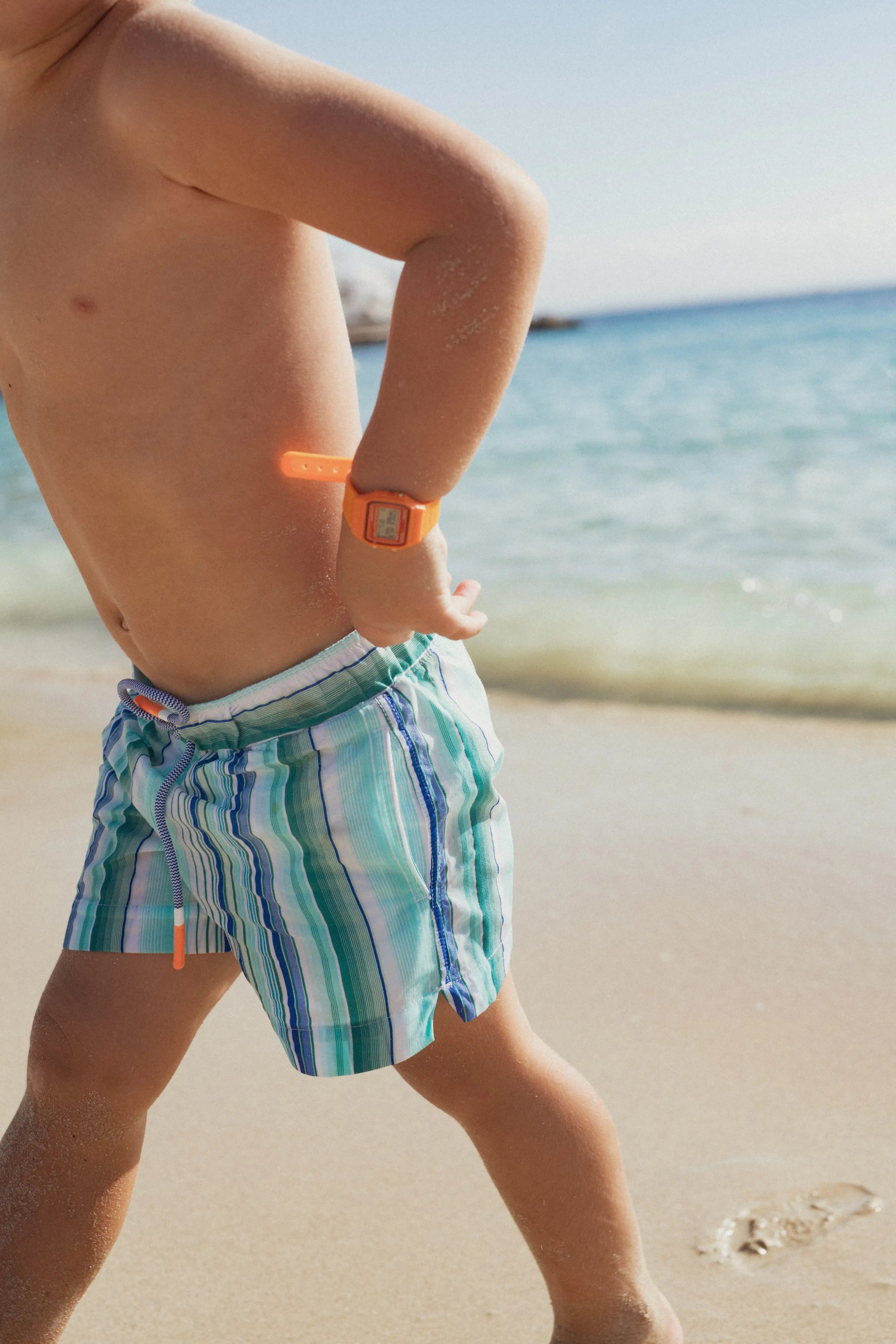 Boy wearing striped swim shorts and orange watch on the beach, Lison Paris swimwear collection.