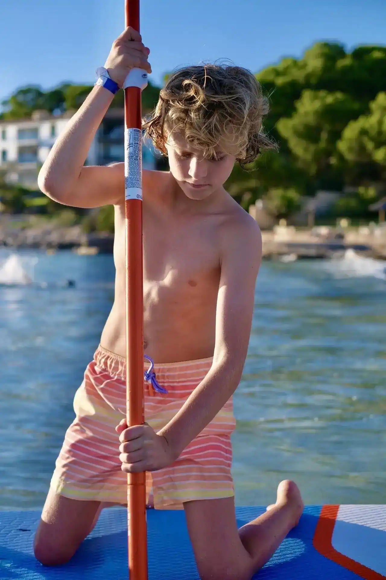 Boy wearing striped swim shorts paddleboarding on the water at the beach