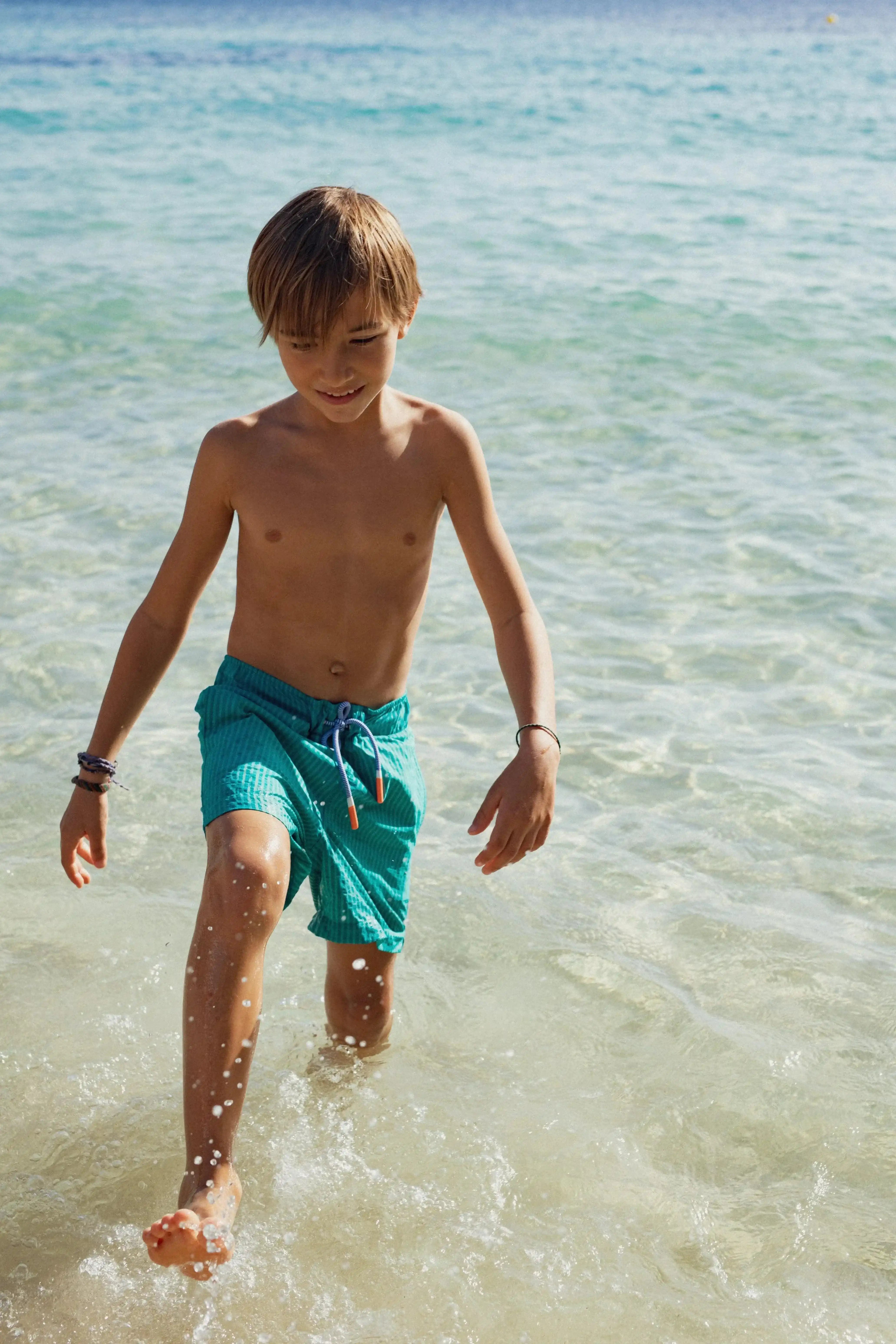 Boy in green striped swim shorts walking in clear beach water, Lison Paris kids beachwear
