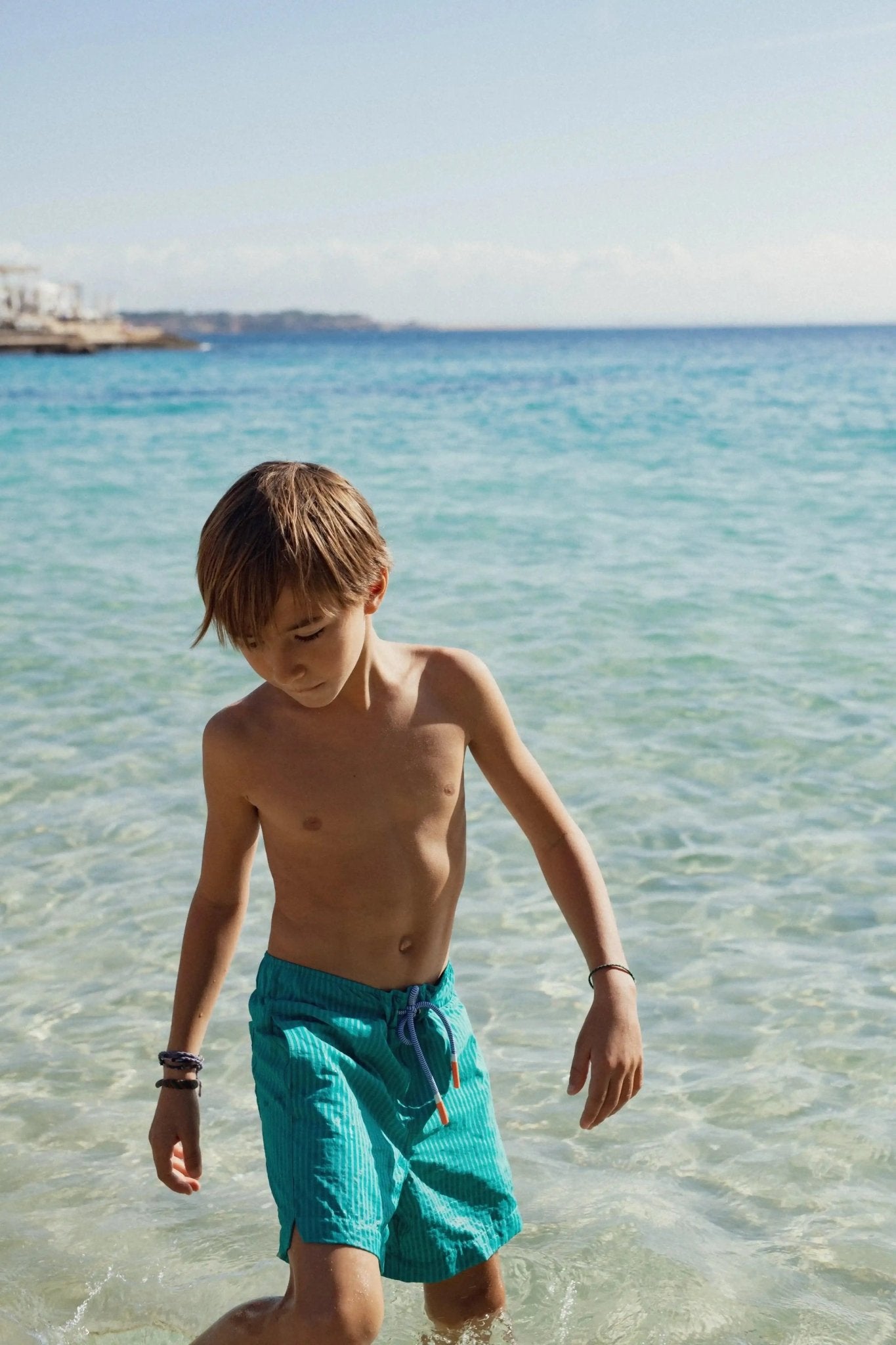 Boy in green striped swim shorts walking in clear shallow sea water on a sunny beach