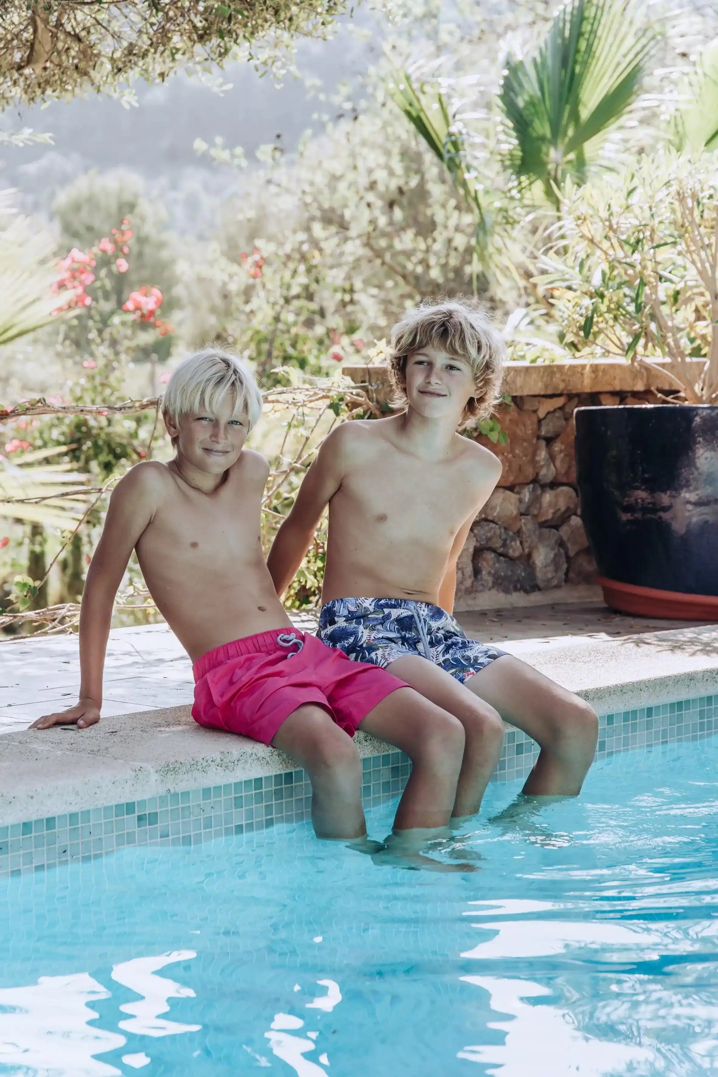 Two boys in swim shorts sitting poolside, one in pink swim trunks, with a sunny garden view