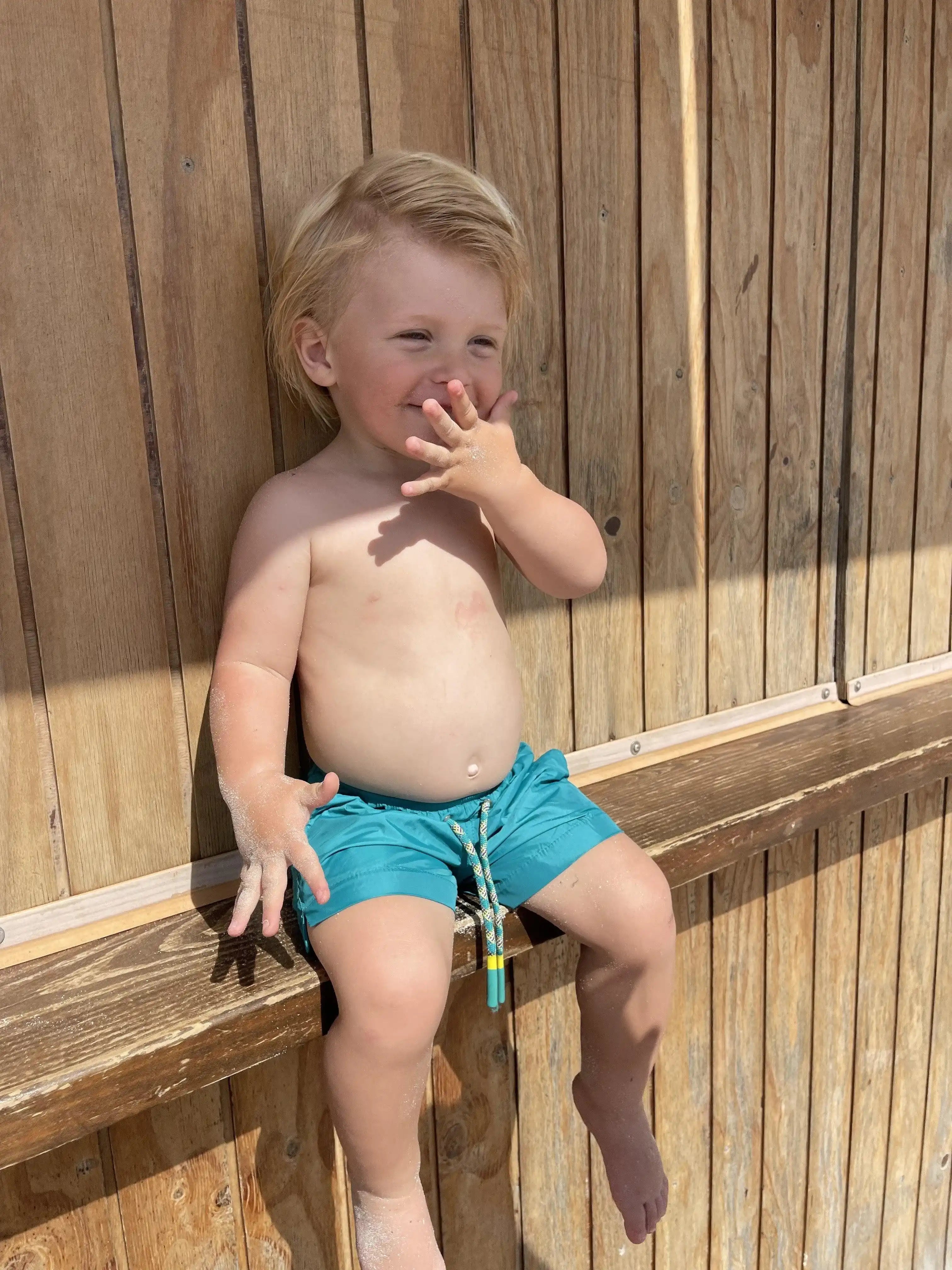 Boy in green swim shorts sitting by a wooden wall, Lison Paris beachwear for boys