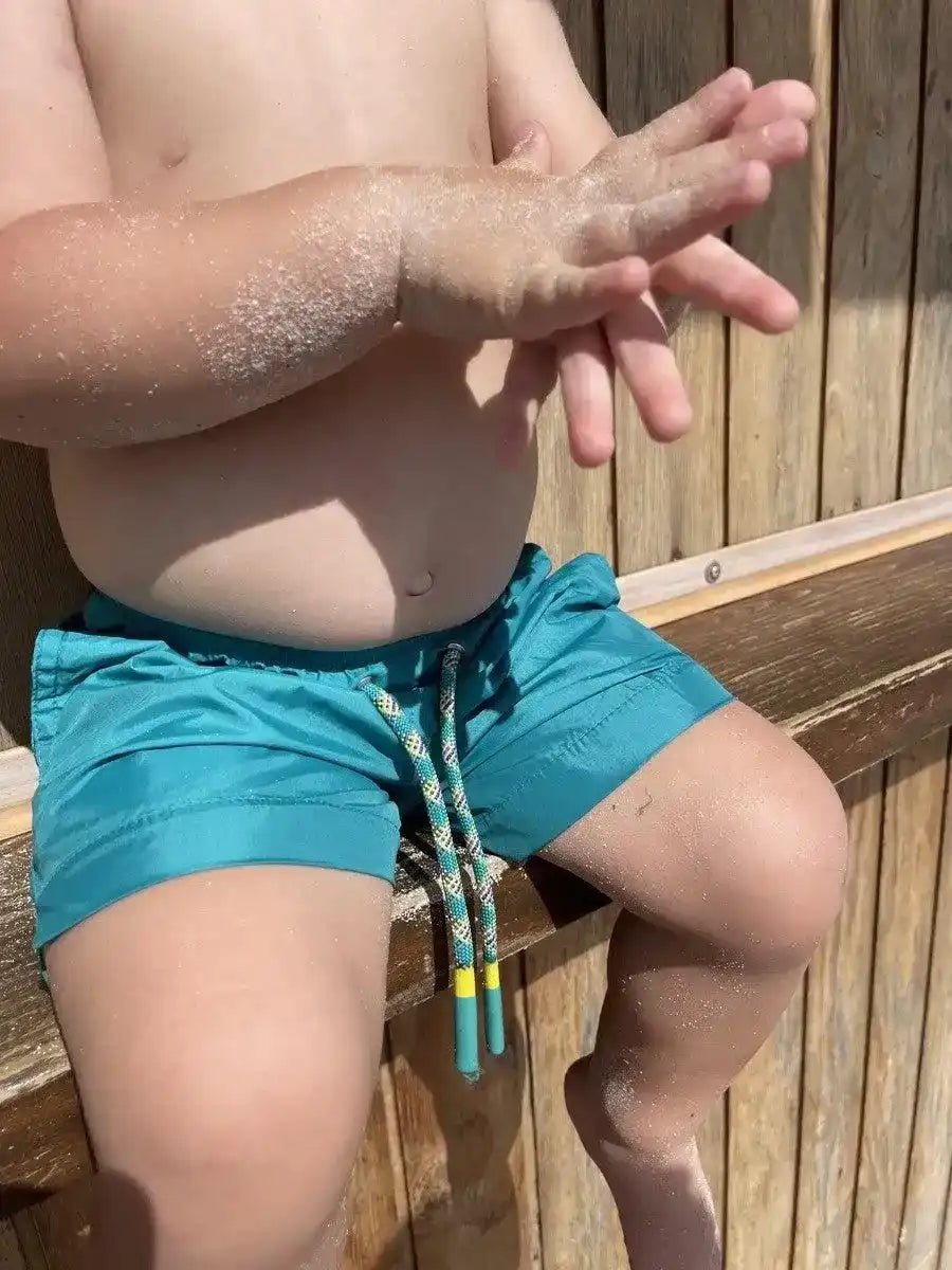 Boy wearing green swim shorts with drawstring, sandy hands, sitting by wooden wall