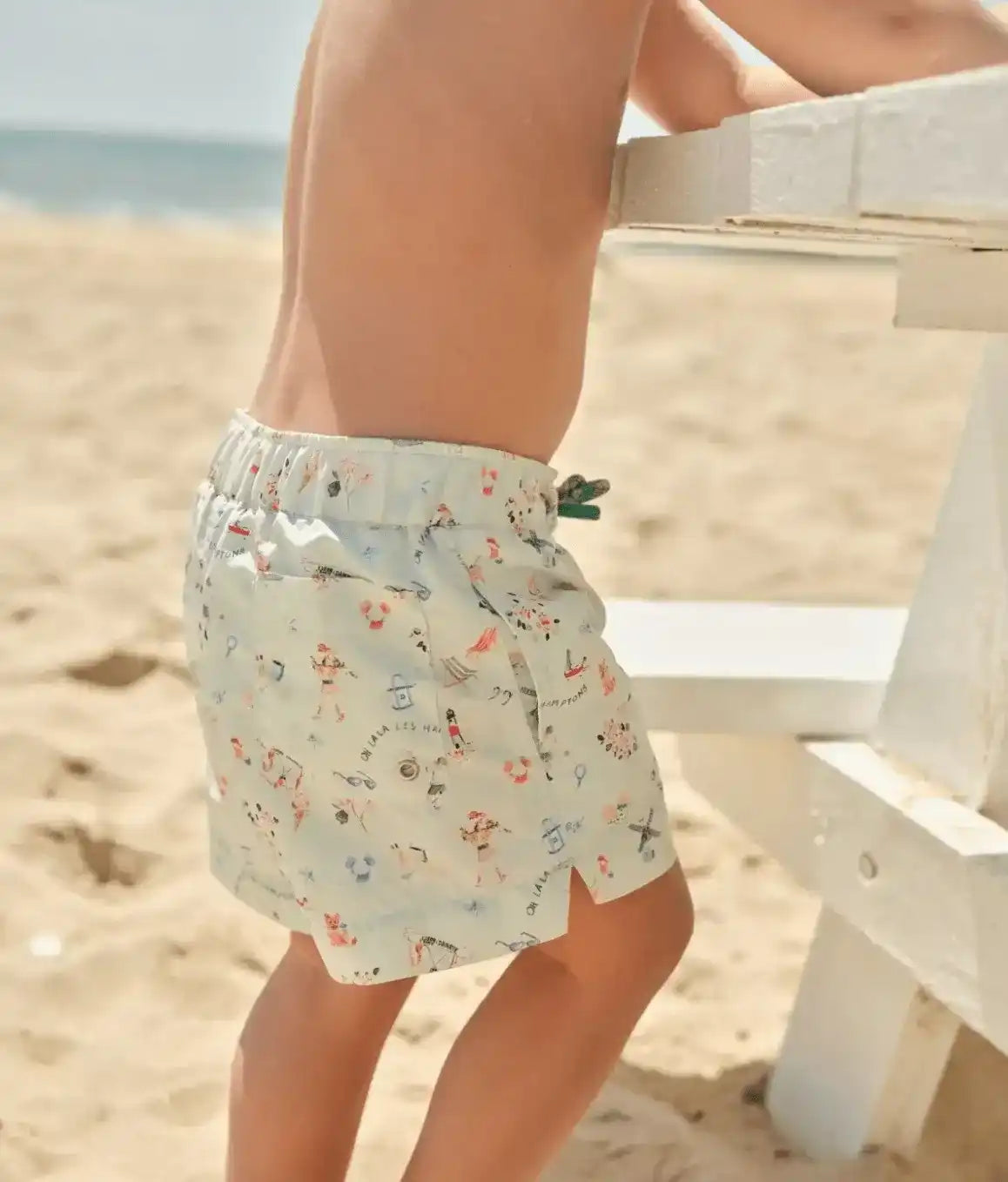 Child wearing printed swim shorts on a sandy beach, leaning on a white bench