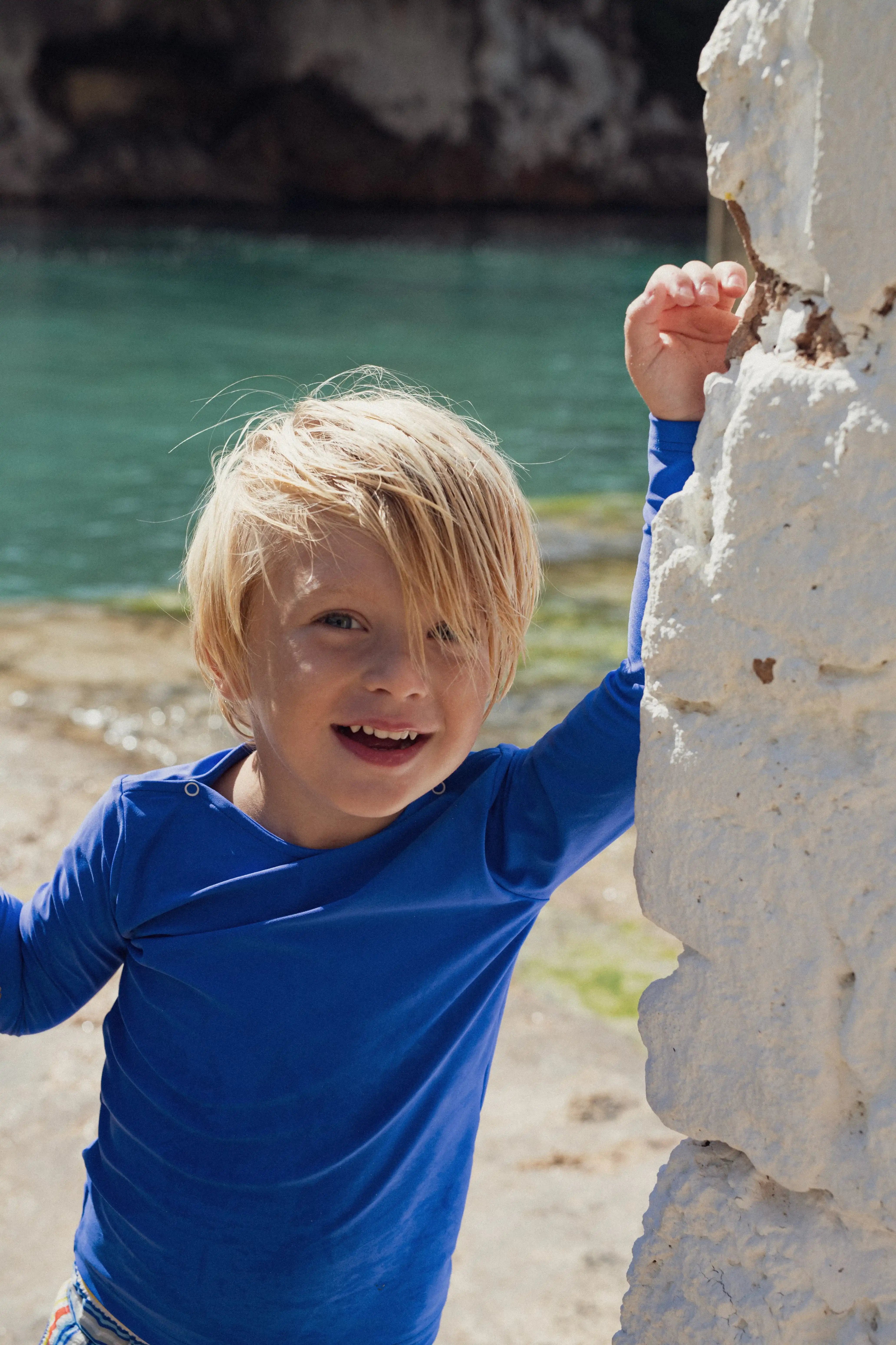 Smiling boy in blue Lison Paris anti-UV swim shirt by the water, beachwear for boys