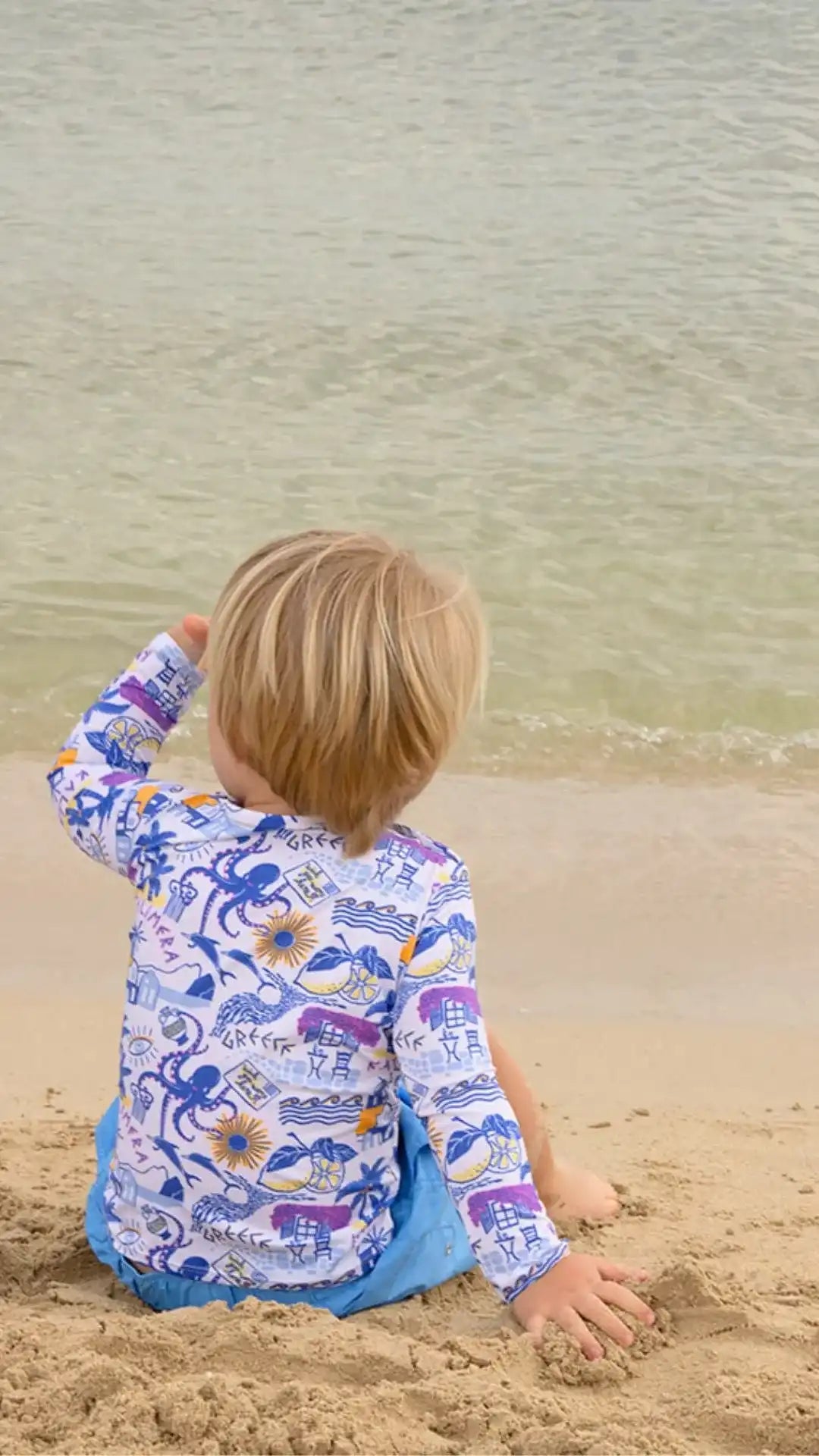 Boy in Lison Paris anti-UV Greek motif shirt sitting on sandy beach by the sea
