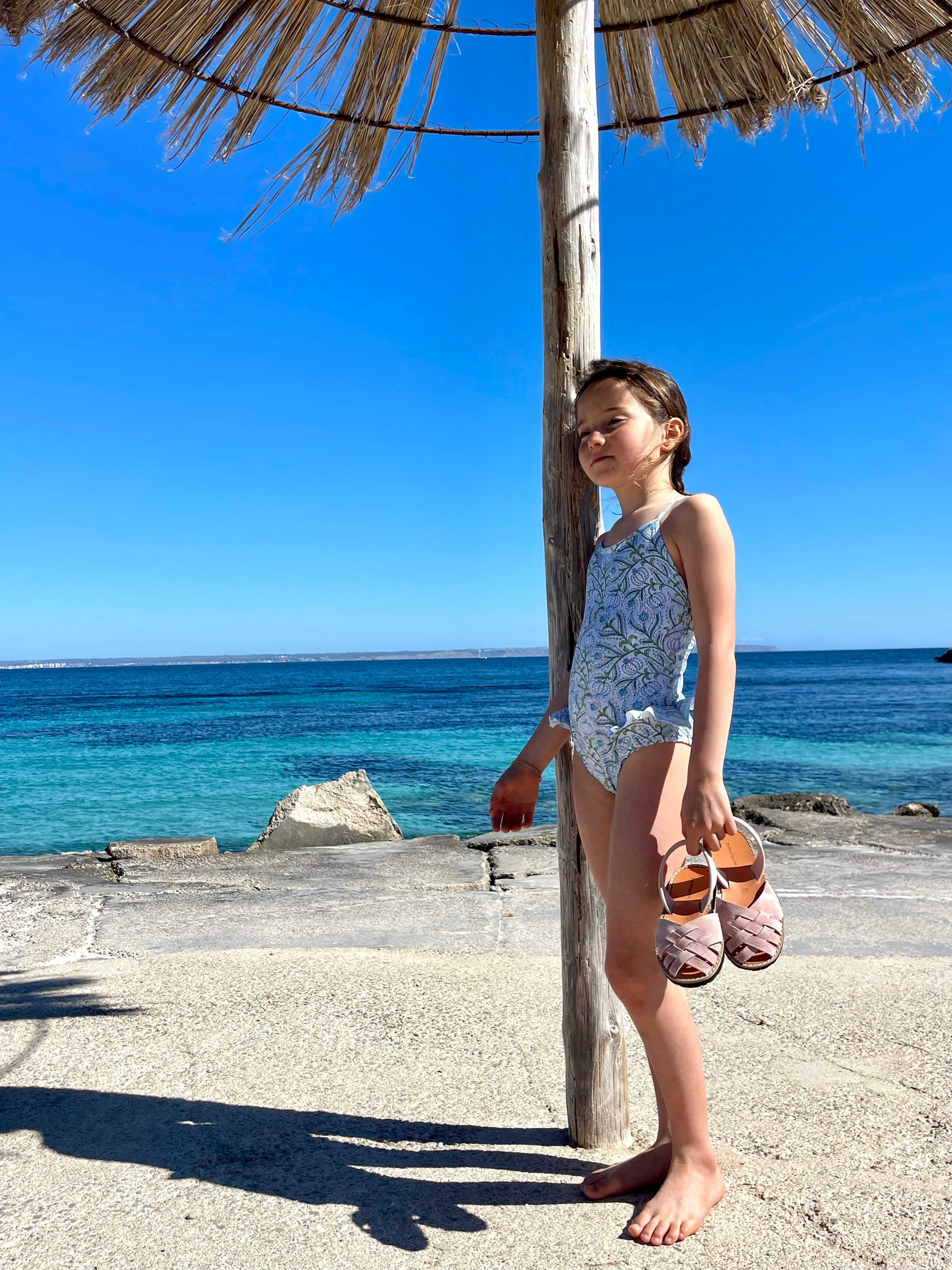 Girl in Lison Paris UV swimwear holding pink sandals, standing by the seaside under a straw parasol