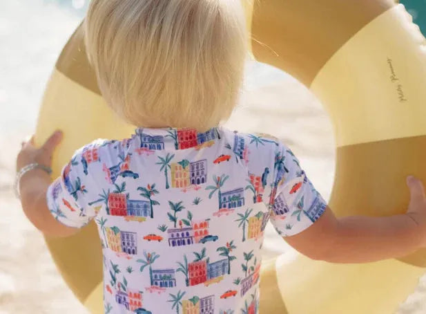 Toddler in colorful printed UV swim shirt holding yellow swim ring on the beach