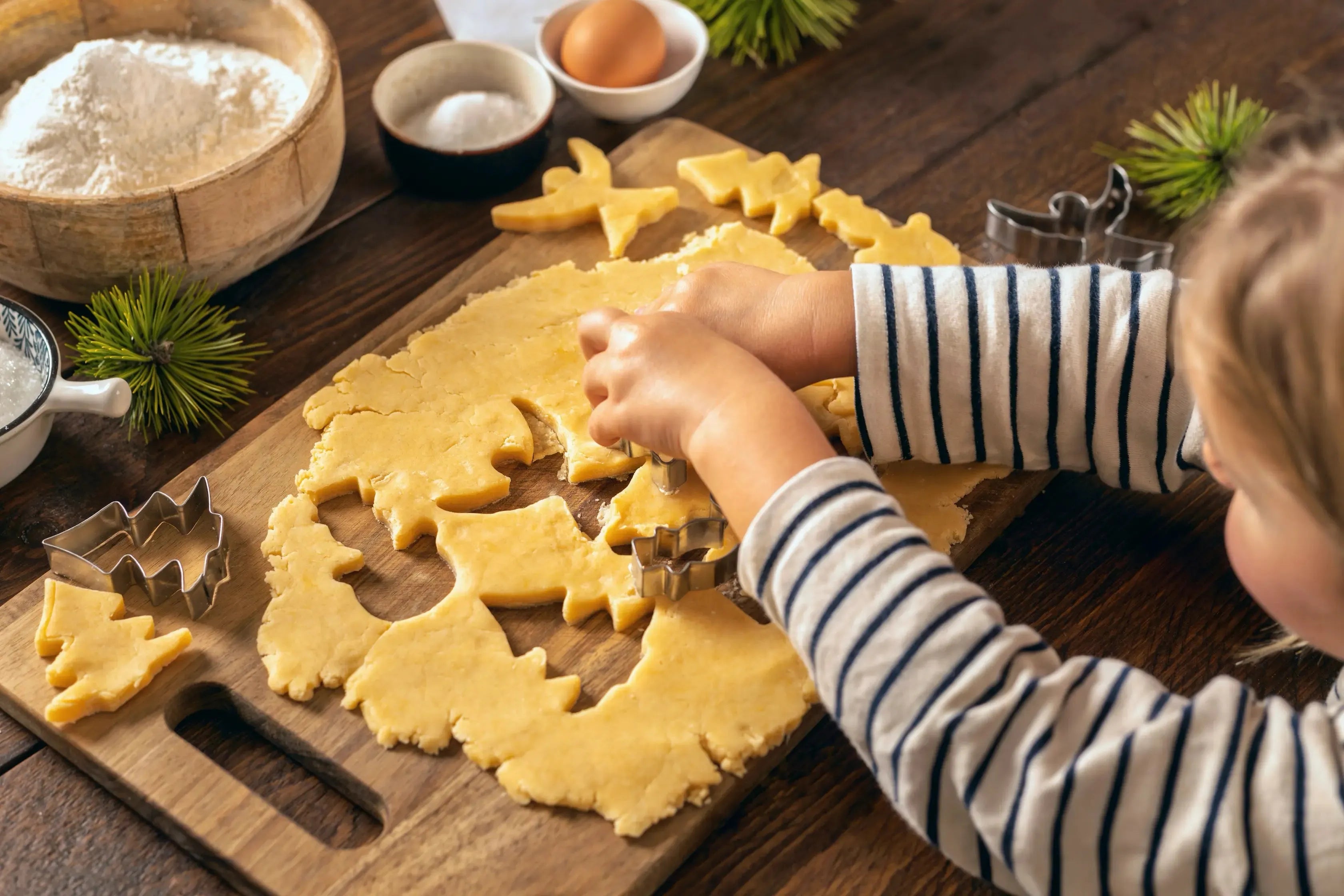 Child cutting Christmas-themed cookie dough with metal cutters on wooden board for holiday baking