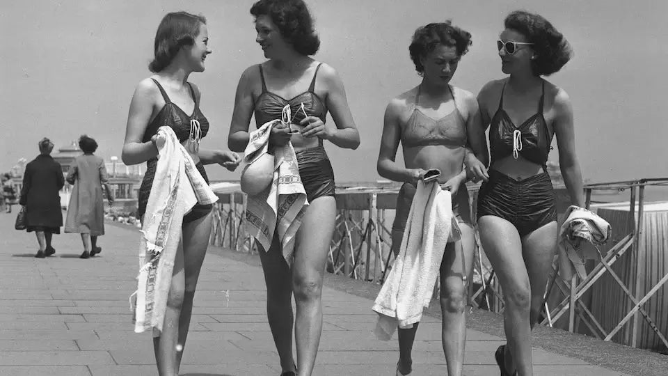 Vintage black and white photo of four women in retro swimsuits walking on a boardwalk, holding towels, with a seaside backdrop.