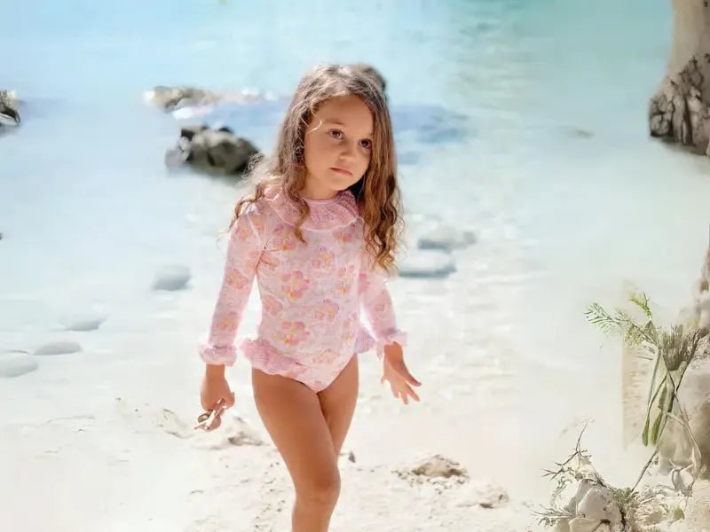 Girl in pink long-sleeve floral swimsuit on sunny beach with clear blue water
