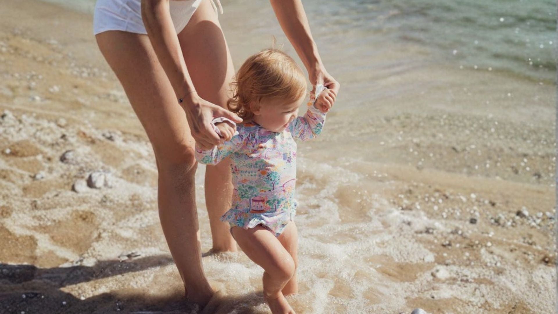 Baby in long-sleeve floral swimsuit playing at sunny beach with adult