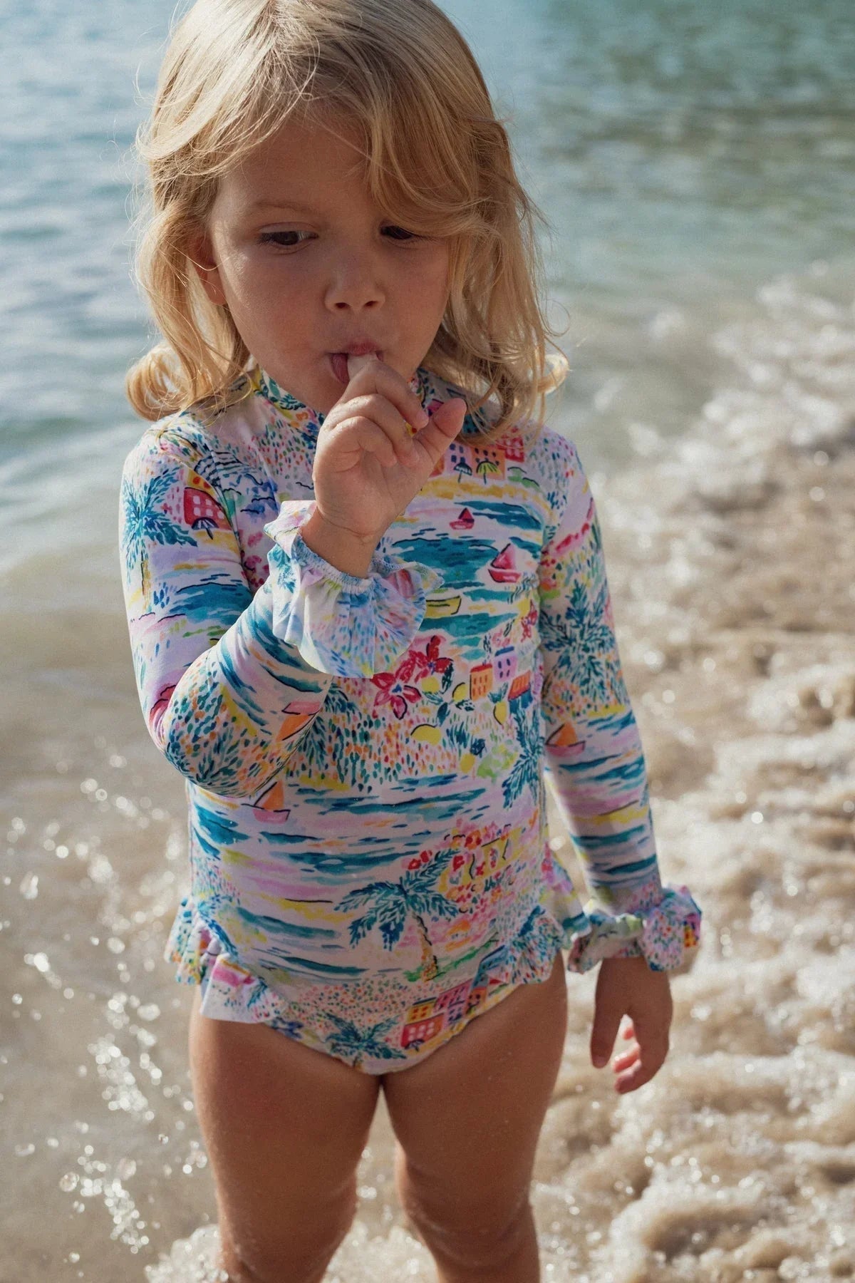 Toddler girl in long sleeve UV swimsuit with colorful print standing on the beach