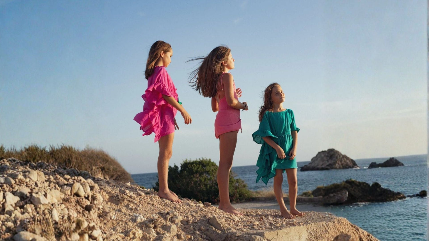 Three girls in colorful beachwear pareos standing barefoot on a rocky seaside cliff at sunset.