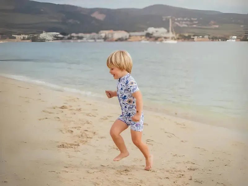 Happy child in anti-UV swimsuit running on sandy beach by the sea, summer vacation mood