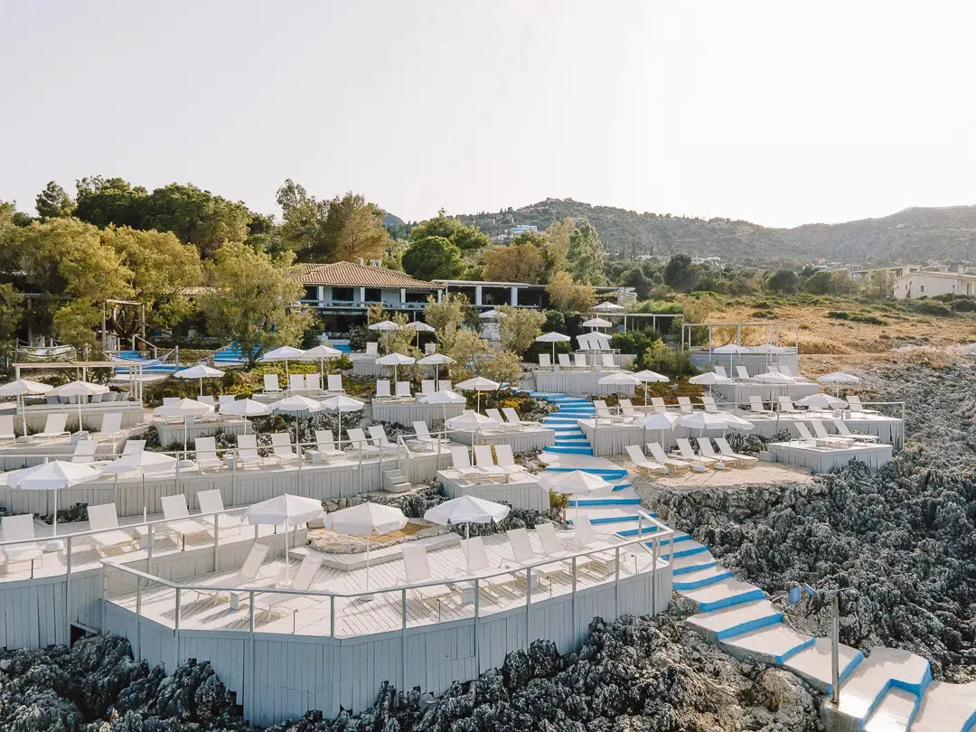 Terraced white wooden beach club with umbrellas and lounge chairs on rocky coastline, surrounded by greenery