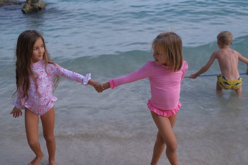 Two girls in long sleeve anti-UV swimsuits playing on the beach, boy in background near water.