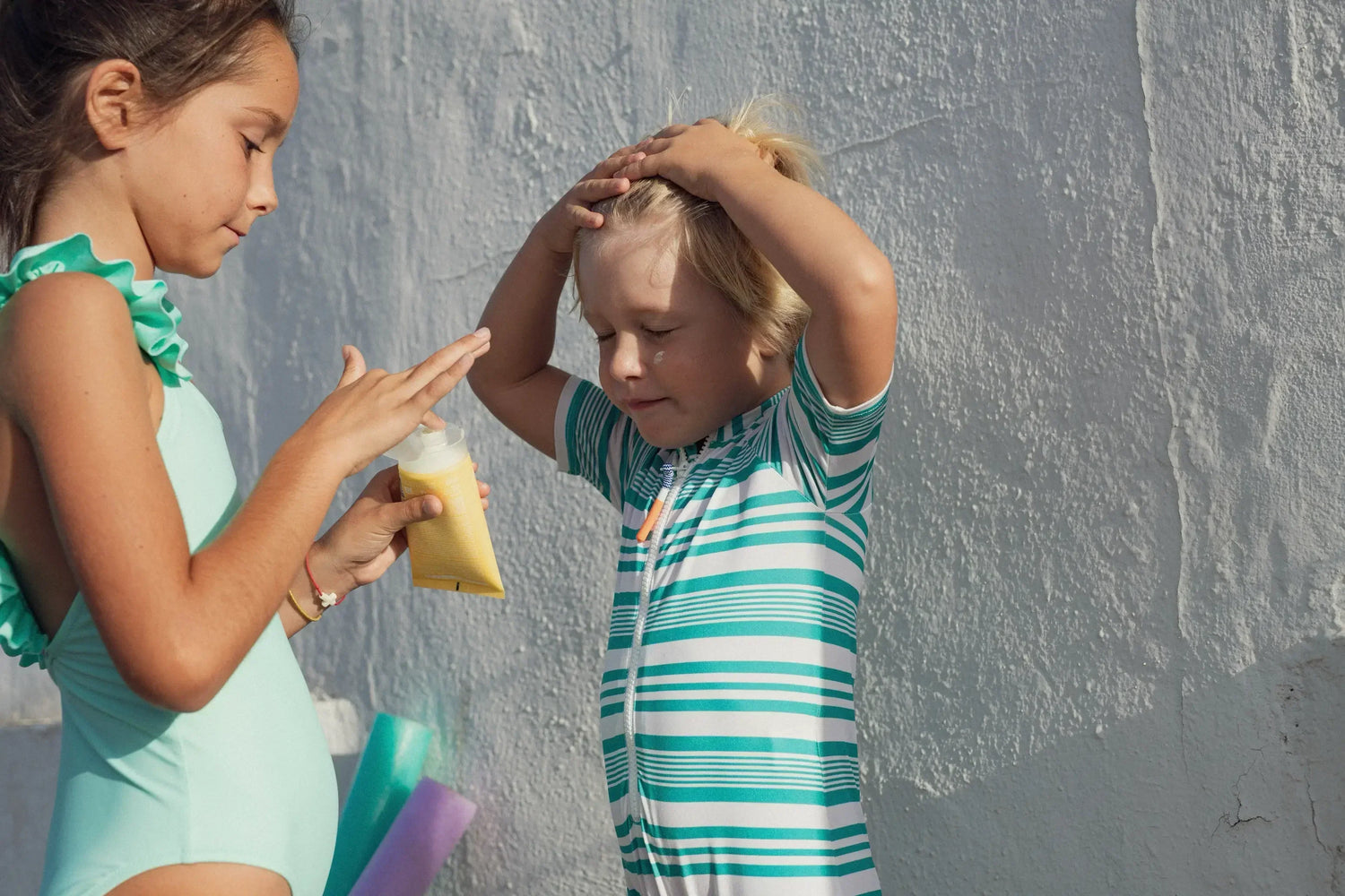 Two children in swimsuits applying sunscreen against a white textured wall on a sunny day