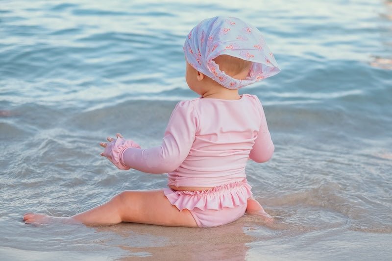 Baby in pink UV-protective swimwear and headscarf sitting at the beach shoreline
