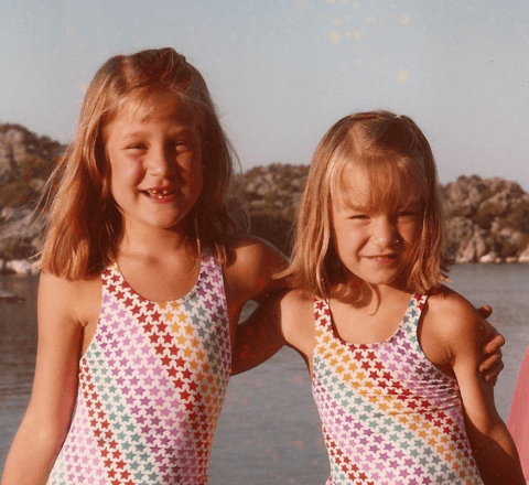 Two young girls smiling outdoors in colorful patterned swimsuits by the water