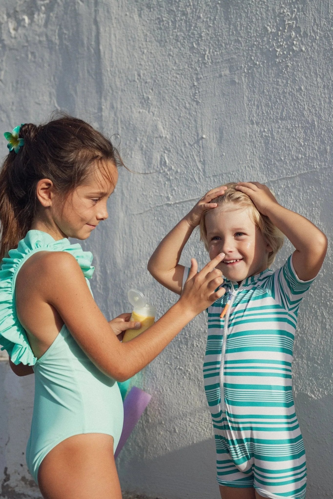 Two children in UV swimwear applying sunscreen outdoors against a sunlit wall
