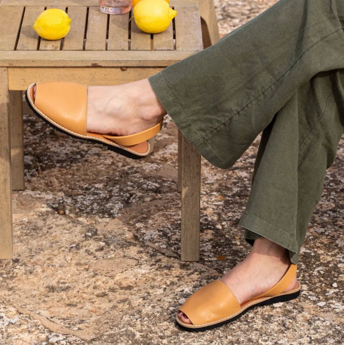 Woman wearing tan leather sandals with open toe, sitting outdoors next to a table with lemons.