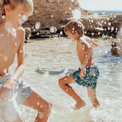 Two boys wearing swim shorts playing in shallow beach water with splashes under the sun