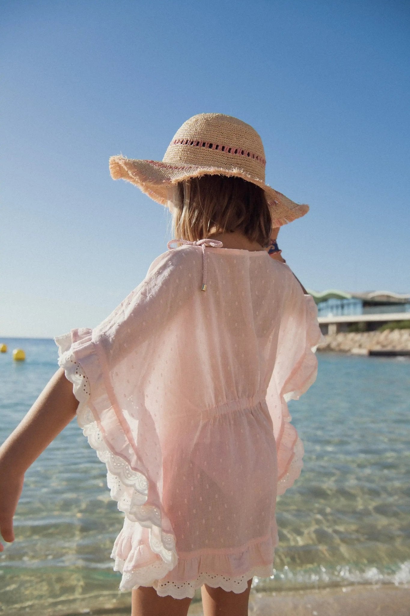 Girl in light pink beach caftan and straw hat standing by the sea on a sunny day