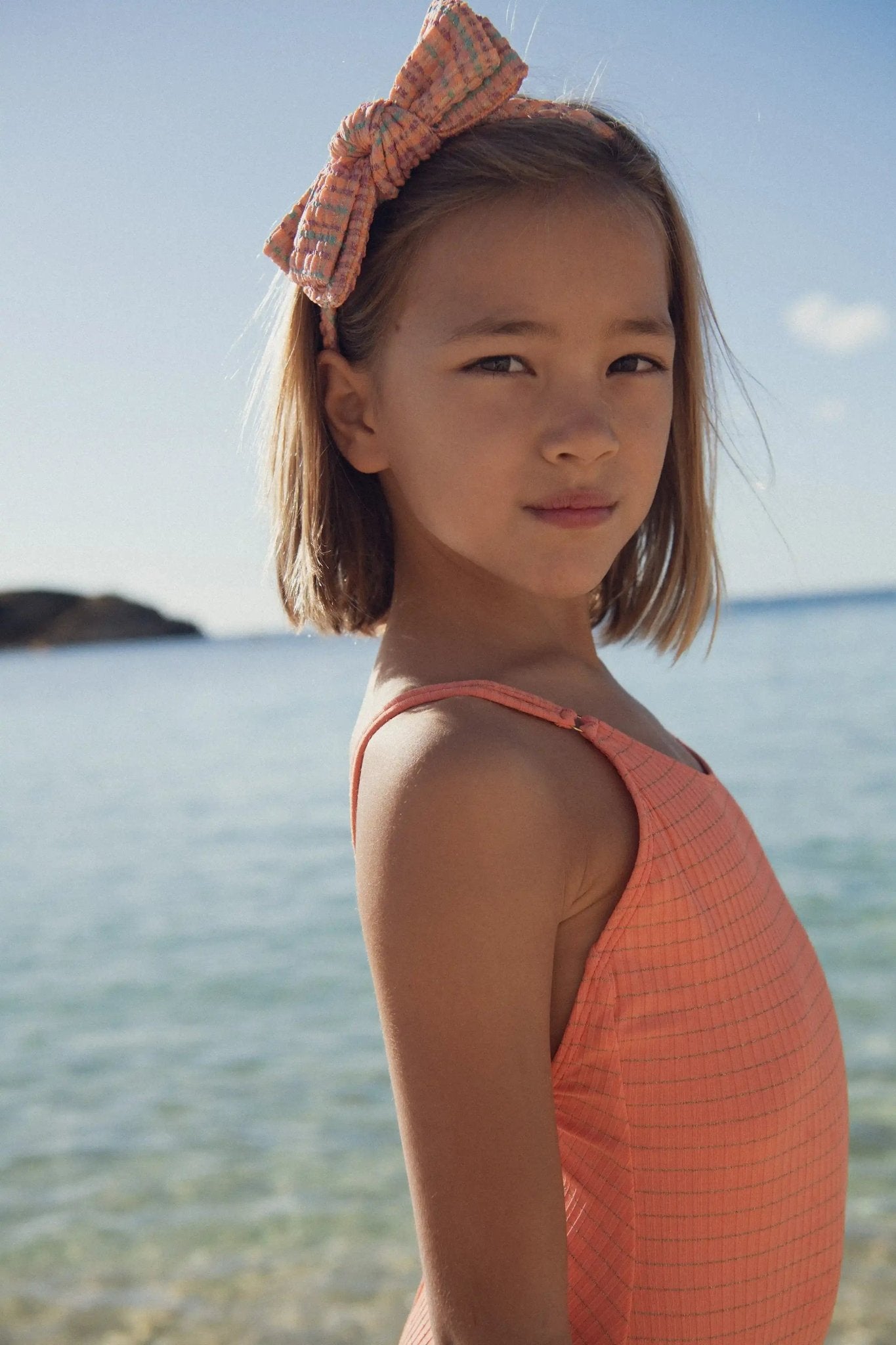 Girl in coral-pattern one-piece swimsuit and bow headband at the beach under sunlight