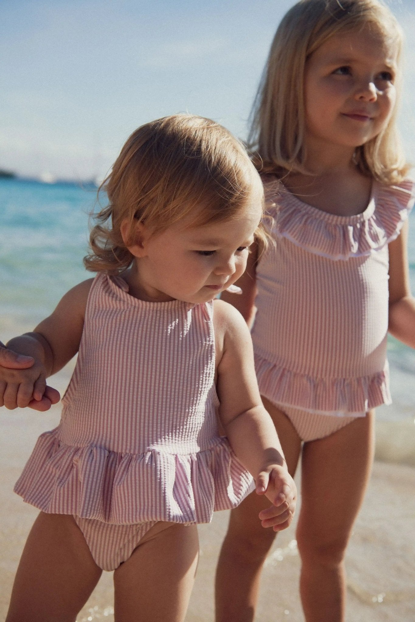 Two young girls in matching pink striped ruffled swimsuits on a sunny beach