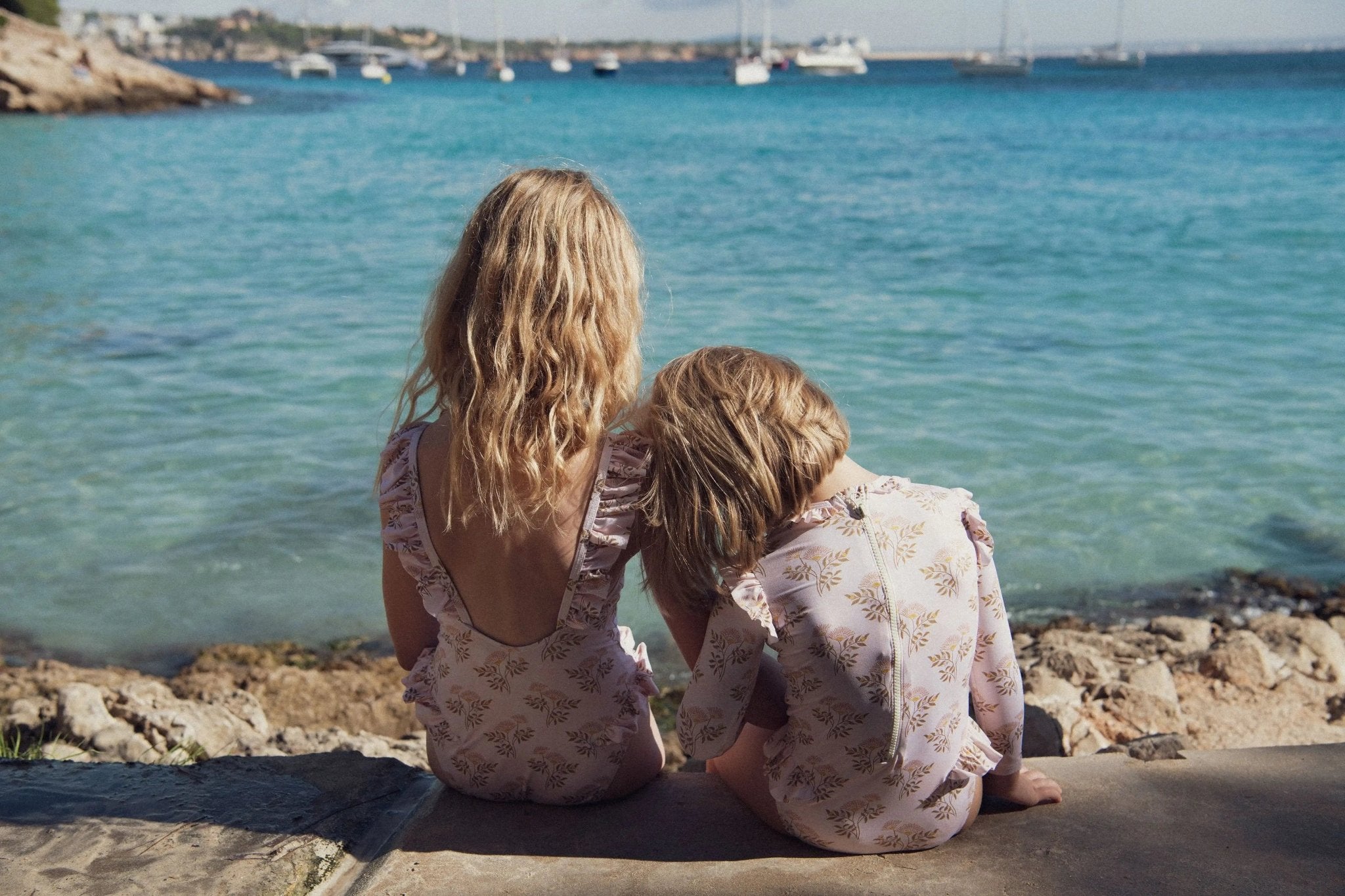 Two children in floral print swimsuits sitting by the sea with boats in the background