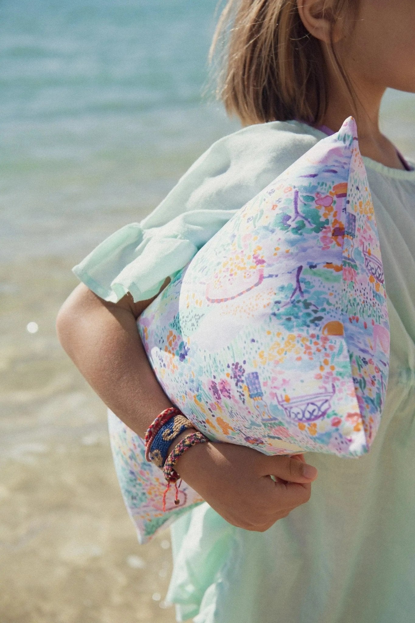 Child at the beach in pastel dress holding a colorful patterned pillow, summer accessories