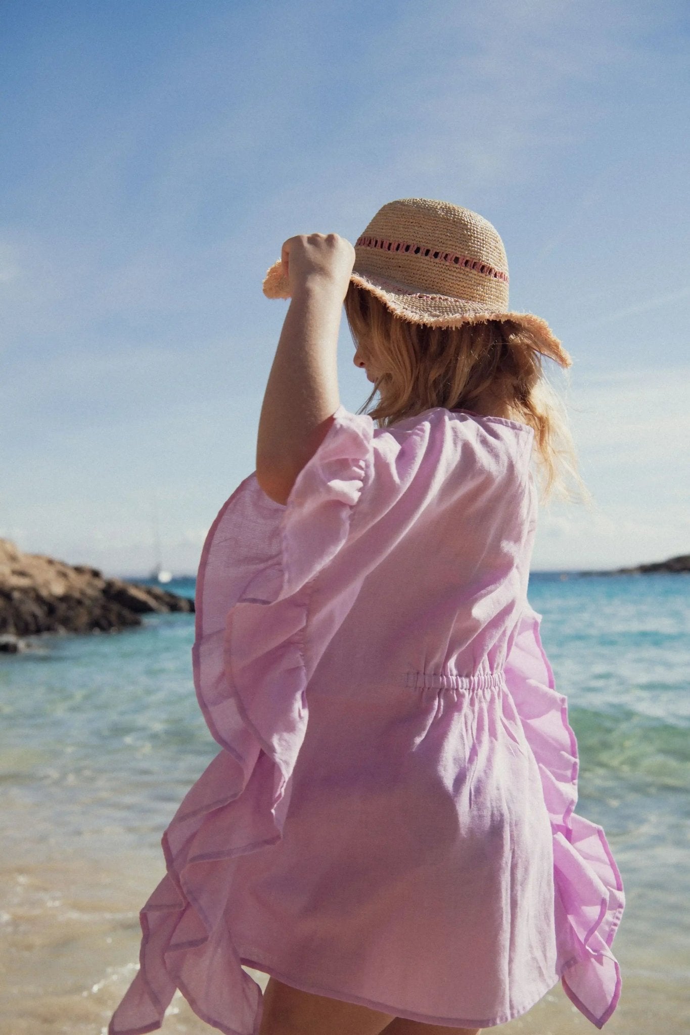 Girl in lilac beach kaftan and straw hat standing by the sea on a sunny day