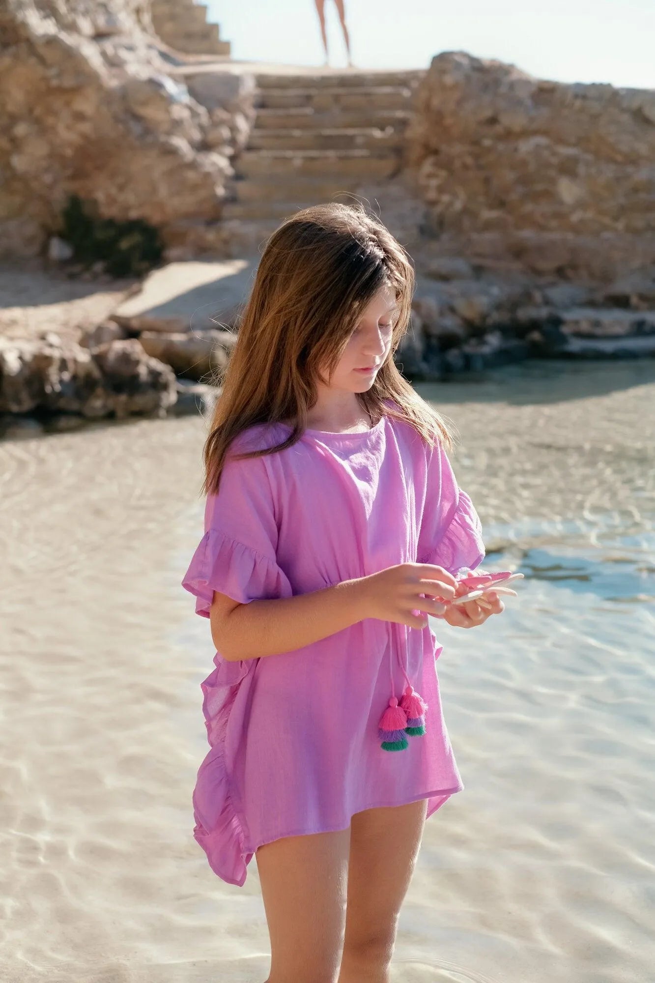 Young girl wearing a pink caftan dress with tassels standing in shallow clear water near rocky steps