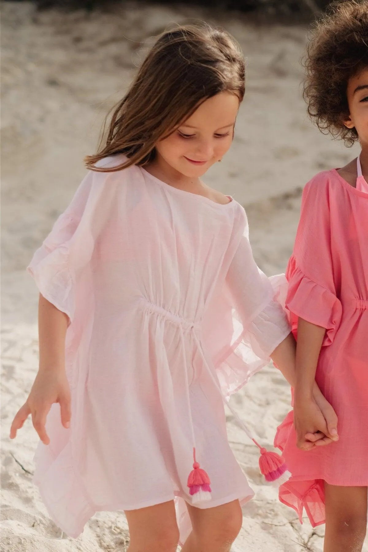 Young girl wearing pale pink pompom caftan dress holding hands with another child on a sandy beach