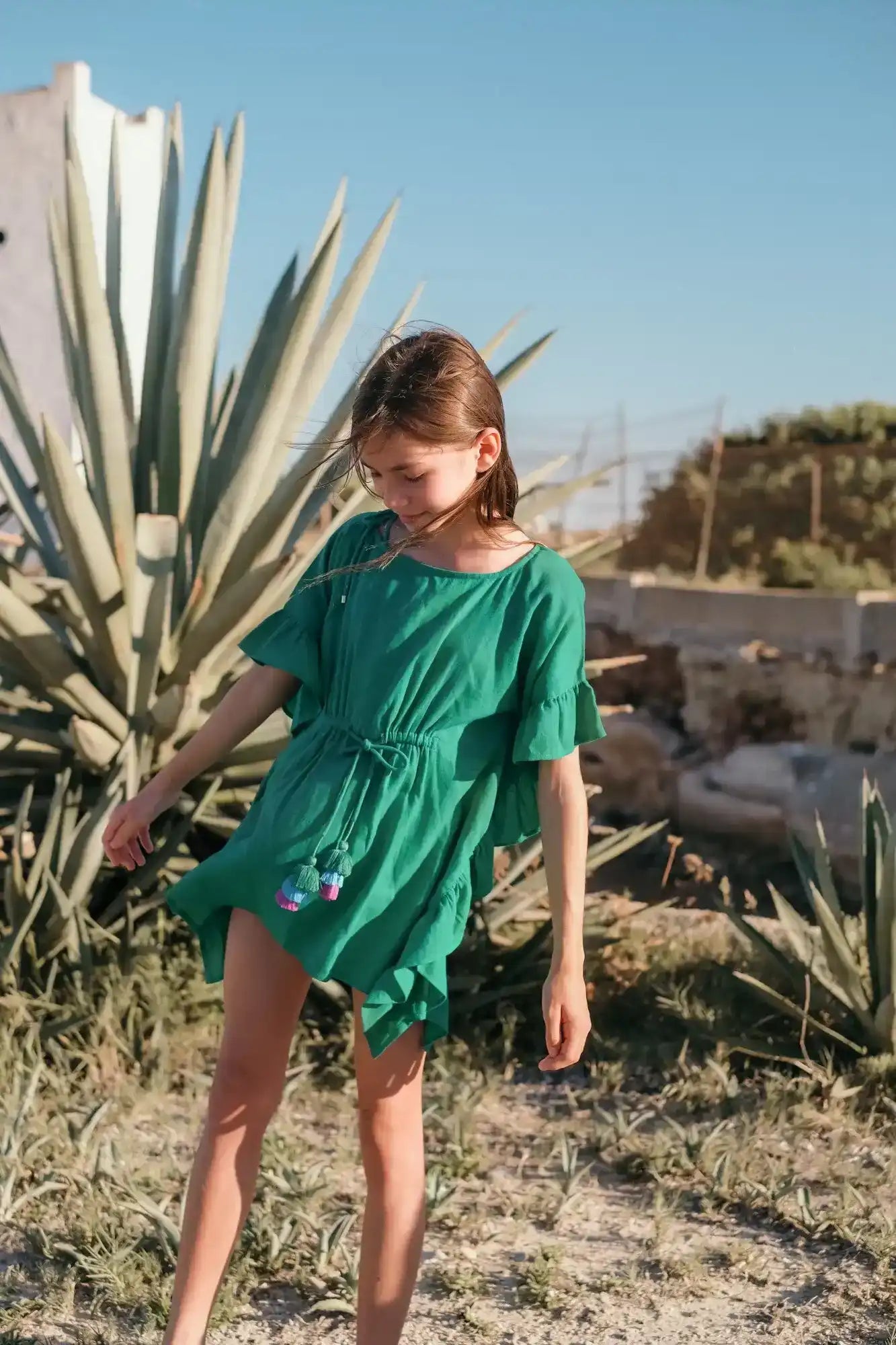 Girl in green caftan with tassel pompoms outdoors, summer beachwear for kids